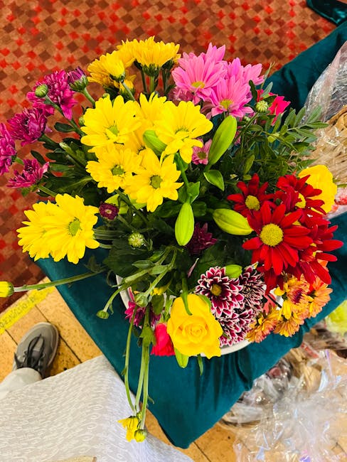 A vibrant floral bouquet featuring large yellow chrysanthemums, bright red daisies, pink chrysanthemums, and purple and white carnations, all with fresh green stems and leaves. The flowers are arrange