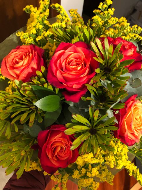 A close-up view of a vibrant floral bouquet featuring large, fully bloomed roses in shades of pink and orange, complemented by sprigs of small yellow flowers and lush green foliage with elongated leav